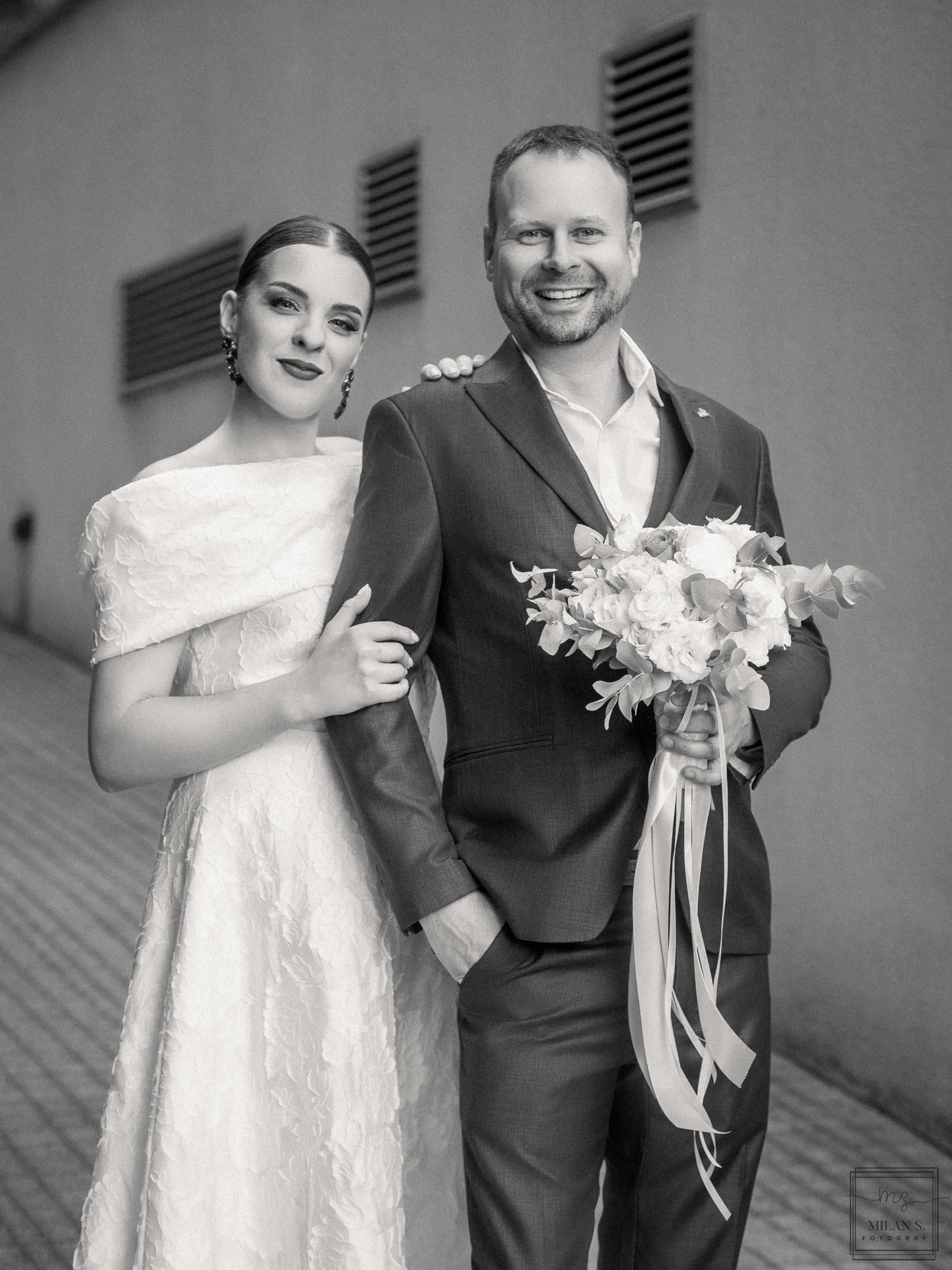 Crno-bela fotografija nasmejanih mladenaca; mladoženja u odelu drži buket sa dugim trakama.