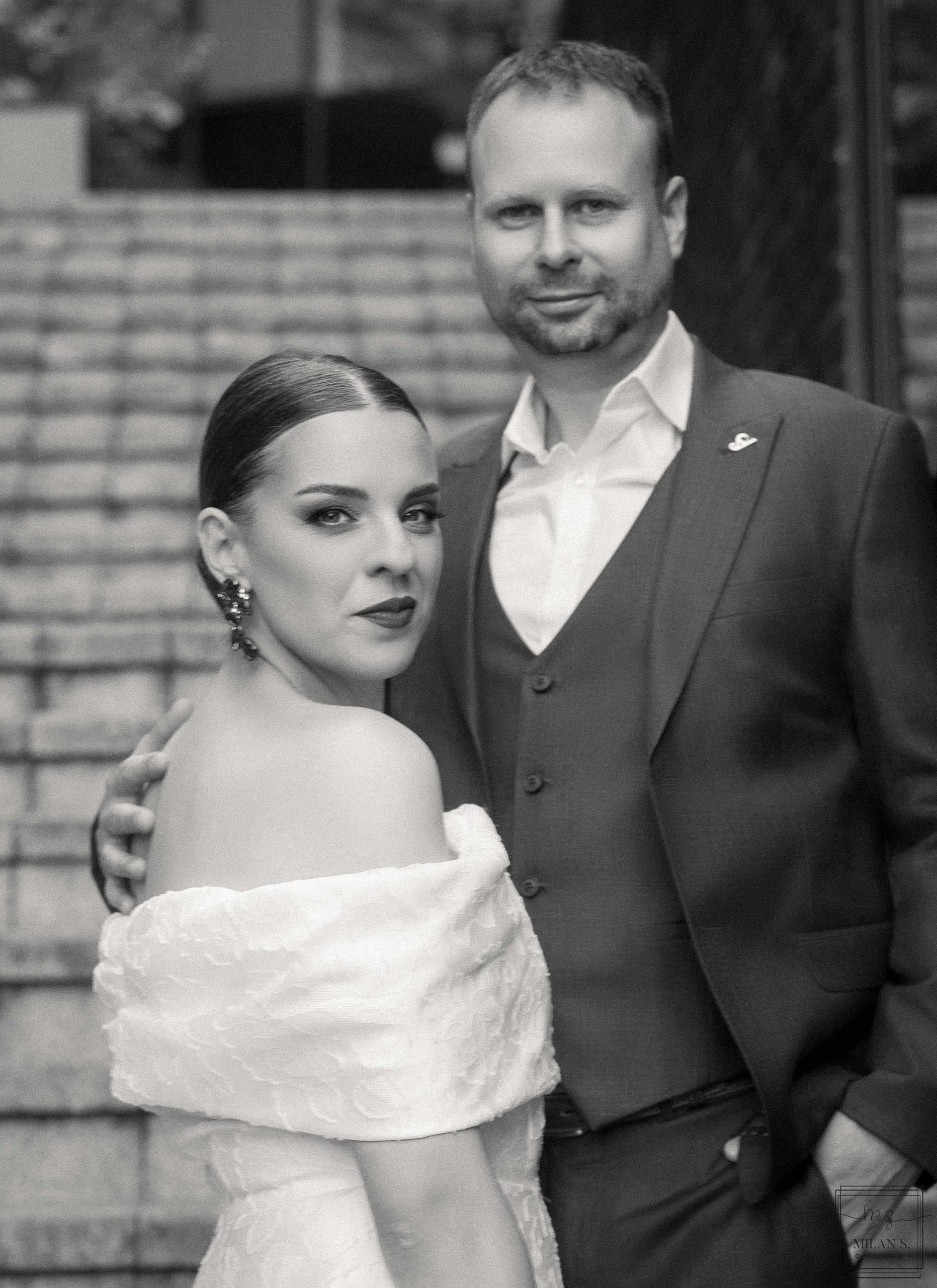Crno-bela fotografija mladenaca na kamenim stepenicama; mlada u haljini bez ramena gleda u kadar.
